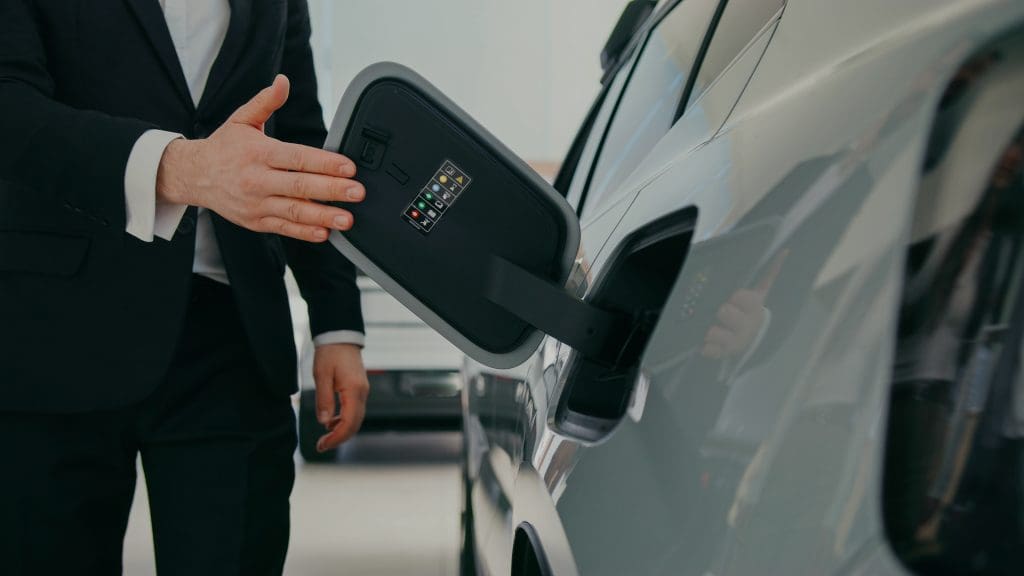 Camera Focus On Person In Black Suit And White Shirt Opening Charging Dock On Side Of Vehicle. Label Printed Inside Of Small Door With Instructions For Future Owners. Car Dealership.