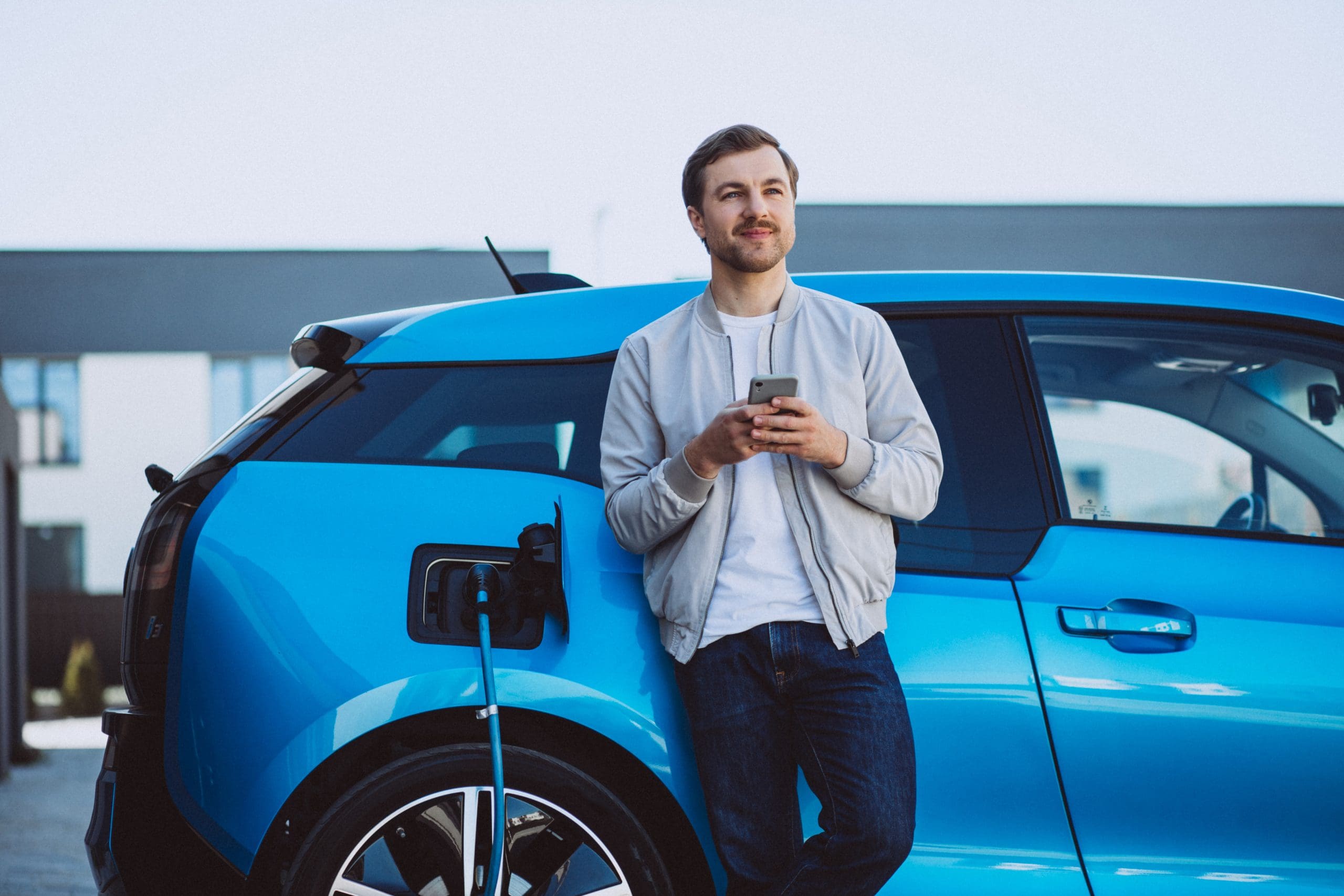Young Man Charging Electric Car At Home And Using Mobile Phone